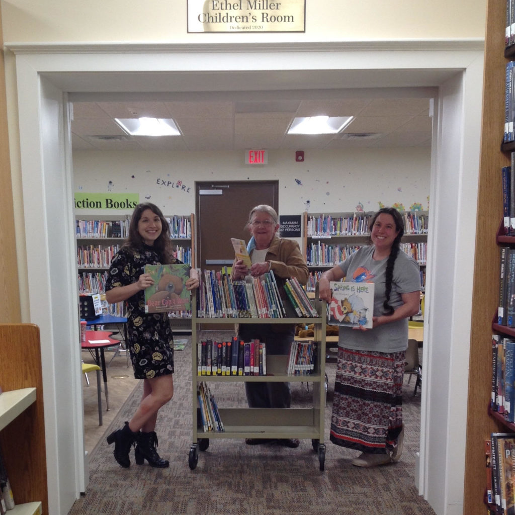 Three women smiling standing with Library cart in a library